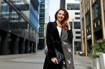 Fashionable beautiful woman walking at the street. Stylish girl dressed in the fur coat and clutch. Female with red hair.