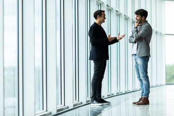 Two businessmen deep in discussion together while standing in an office boardroom with windows overlooking the city