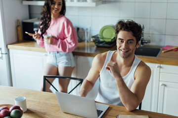 Selective focus of smiling man using laptop near girlfriend with cup and smartphone in kitchen