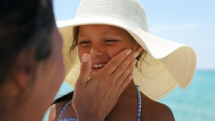 Close up of young mother is applying protective sunscreen or sunblock lotion on her little happy smiling daughter's face to take care of skin on a seaside beach during family holidays vacation. - Powered by Adobe