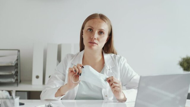 Beautiful female doctor in lab coat sitting at desk in clinic, explaining and demonstrating how to wear medical face mask at camera during covid-19 outbreak