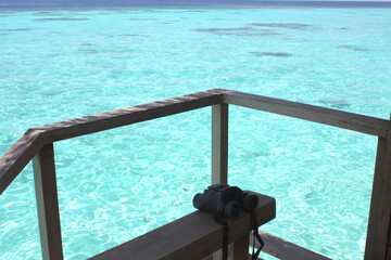 Fototapeta na wymiar Beautiful isolated black binoculars on an overwater bungalow, with the beautiful turquoise blue sea of a lagoon in Indian ocean in background, Maldives Island.