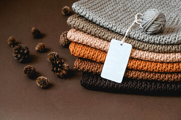 Stack of knitted material from threads of brown, orange, gray colors with an empty price tag on a brown background. Copy, empty space for text