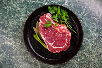 Fresh Raw Meat for Steak Ribeye, Decorated with Herbs and Sprinkled with Spices and Coarse Salt, Lies in a Cast-Iron Vintage Pan. View from Above. Green Marble Table.