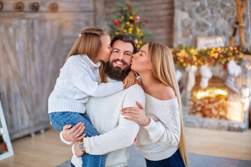 Fototapeta premium Family Christmas concept. Lovely family in bright clothes standing near decorated Xmas tree on winter evening. Happy household with daughter on floor at home near fireplace