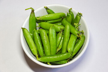 Peas in a pod.  Freshly picked sugar snap peas in a bowl.  The vibrant green pea pods are set against a white background.  These high fiber, low calorie vegetables are ideal for a healthy snack.