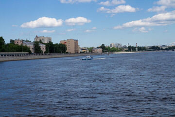 Boat on a wide river, against the backdrop of the city landscape and blue sky with clouds.