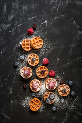 Obraz na płótnie Canvas Mini waffles with raspberries and blueberries on a black stone background, flat lay. Belgian traditional waffles with berries. Top view, copy space.