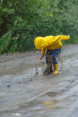 Kid playing in the rain in autumn park. Child jumping in muddy puddle on rainy fall day. Little boy in rain boots and yellow jacket outdoors in heavy shower. Kids waterproof footwear and coat. 