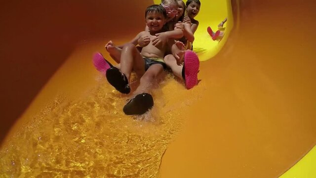 Group of happy children riding down an orange slide in a water park. Children laugh merrily and wave their hands