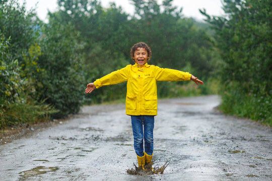 Kid Playing In The Rain In Autumn Park. Child Jumping In Muddy Puddle On Rainy Fall Day. Little Boy In Rain Boots And Yellow Jacket Outdoors In Heavy Shower. Kids Waterproof Footwear And Coat. 