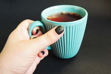 a woman's hand with black nails holds a turquoise textured mug with fruit tea on a black background