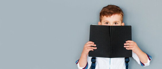 Childhood and education concept - little boy hiding behind book over grey background