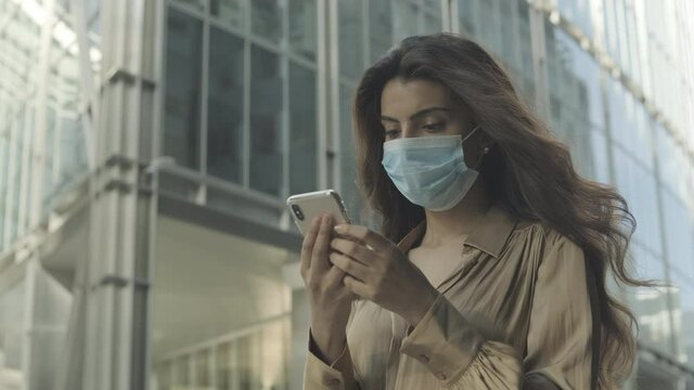 Young adult business woman commuting in city wearing protective face mask and using mobile phone on the move during pandemic