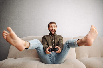 A joyful young man enthusiastically plays on a console, sitting on the couch holds a game...