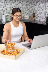 Asian woman work from home in the kitchen and eating delivery pizza take away food and take out coffee while city lockdown from coronavirus covid-19 pandemic. New Normal lifestyle working from home.