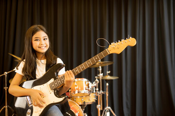 A portrait of a cute Asian girl with long hair playing an electric guitar in a music practice room.