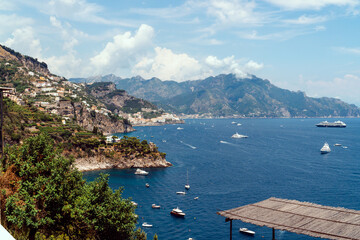 Obraz premium View from Conca dei Marini on Amalfi bay, Luxury boats traffic, ocean liner, fishing boats, tourist season. watercraft. Mountains and houses, buildings. Summer day. Amalfitana Italy