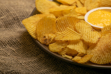 Fluted potato chips in a round plate with sauce in a saucepan in the middle of the plate on a background of homespun fabric with a rough texture. Close up.