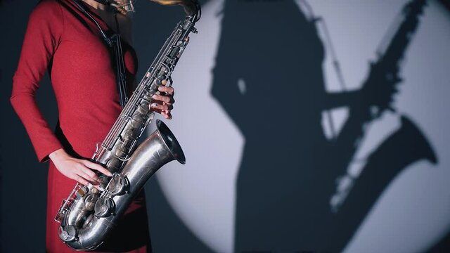 Young beautiful woman in red concert dress playing a melody on saxophone over dark background in the spotlight