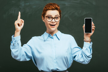 cheerful teacher in eyeglasses holding smartphone with blank screen near chalkboard