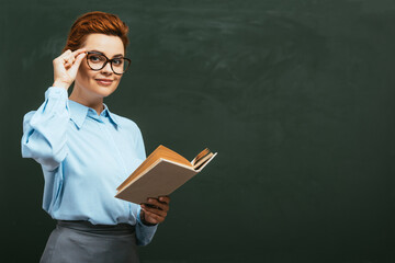 smiling teacher touching eyeglasses while standing near chalkboard with open book