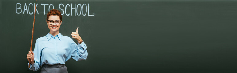 panoramic concept of smiling teacher with pointing stick showing thumb up near chalkboard with back to school lettering