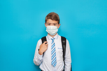 A schoolboy boy wearing a medical mask, shirt and tie with a school backpack on a blue background....