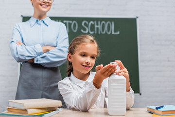 partial view of teacher standing with crossed arms near schoolgirl applying antiseptic on hands