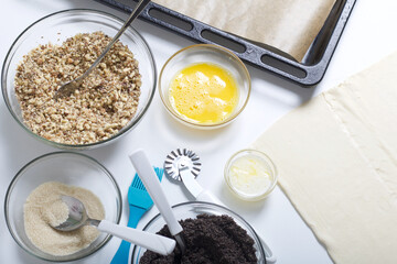 Ingredients and tools for making puff pastry curls stuffed with poppy and walnut. Are laid out on a white table top.