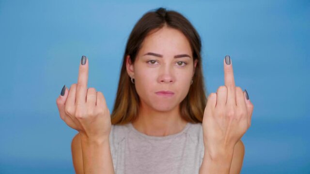 Serious woman in gray t-shirt shows middle finger on blue background with copy space. 4K slow motion footage