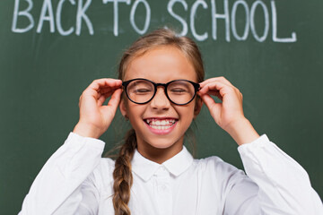 selective focus of excited schoolgirl laughing with closed eyes and touching eyeglasses near chalkboard with back to school lettering