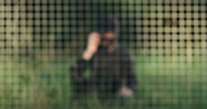 A man in a black mask threatens by phone while sitting on the grass behind bars, blurred background.