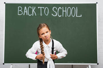 displeased schoolgirl with heavy backpack near chalkboard with back to school lettering