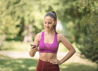 Fototapeta premium Girl athlete in a red uniform smiling in the park with a phone and headphones. Stands right up close.