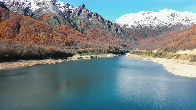 paisaje de un lago o embalse azul con cerros y monta&ntilde;as con nieve de fondo , arboles de color verde y naranjo 