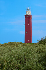 Red lighthouse, square tower, in front of green bushes, blue sky in the background. Ouddorp, Netherlands.