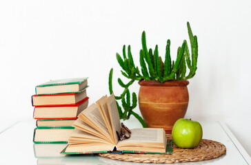 Cozy home interior decor: stack of books, plants in pots, open book and green apple on a wicker stand, pillows on a white table. Distance home education.Quarantine concept of stay home.