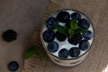 Cottage cheese dessert with blueberries, blackberries and mint in a transparent glass glass. On a burlap napkin and on a wooden background. Delishious.