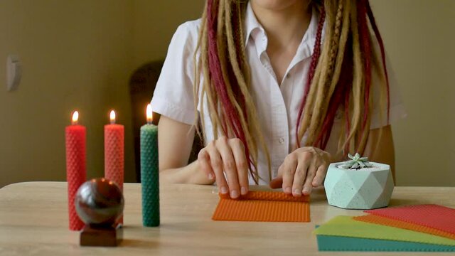 Young modern girl with dreadlocks in white shirt is performing a workshop of making beeswax candles sitting in front the table with stone and plants on it