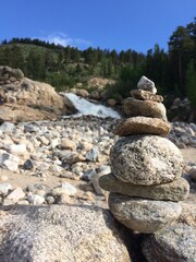 Rock Cairn Stacked Near Mountain Stream with Rocky Beach, Evergreens, Waterfall, and Blue Sky in Background