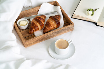 Breakfast in bed with two croissants and butter on the wooden tray, a cup of coffee and open book on the white sheet with blanket. Slow life.