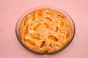A large, round baked apple and apricot pie stands in a glass form on a table with a tablecloth.