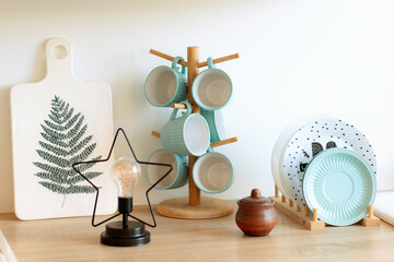 on the kitchen table, a stand with plates and cups for tea. Kitchen wood board. Decorative lamp.
