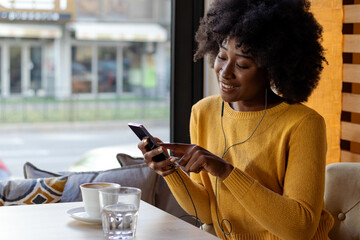 Beautiful black girl with curly hair at cafe table listening to music on smartphone and enjoing in a cup of coffee. Leisure, technology and urban lifestyle concept.