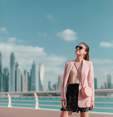Young stylish woman in pink jacket and sunglasses stands on the promenade. Skyscrapers in the background.