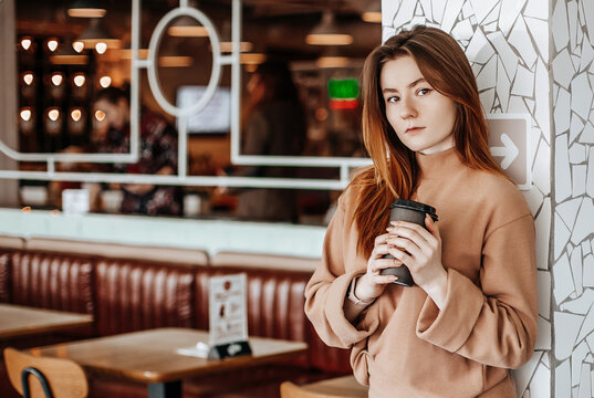 Stylish girl is drinking coffee in a cafe. A woman with ginger hair is standing against a wall in a beige suit. Modern interior. Coffee to go in a cardboard cup