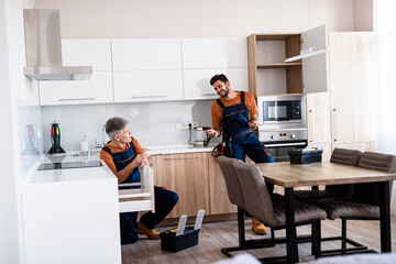 Obraz premium Full length shot of two handymen, workers in uniform talking while assembling kitchen cabinet using screwdriver indoors. Furniture repair and assembly concept