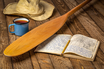 Vintage expedition journal on a rustic picnic table with a cup of tea, canoe paddle and hat. Travel...