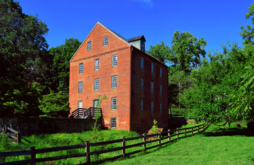 Old historic brick mill building in Waterford Virginia. Green grass field with split rail fence and trees. Summer day with blue sky.  Colonial architecture.